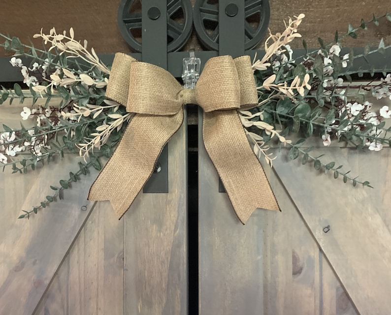 May include: A rustic burlap bow with faux greenery and white flowers, attached to wooden barn doors with metal hardware. The design has a farmhouse aesthetic. The doors are painted in a light grey colour, with a natural wood grain.