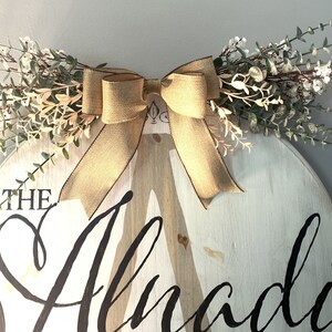 May include: A round, white wooden sign with the name "Alnadi" in black script. The sign is decorated with a burlap bow and greenery. The sign is approximately 46 cm in diameter.