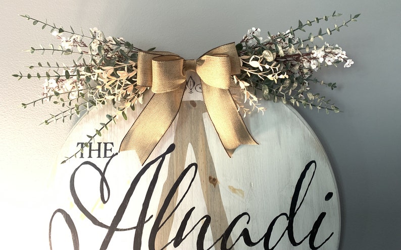 May include: A round, white wooden sign with the name "Alnadi" in black script. The sign is decorated with a burlap bow and greenery. The sign is approximately 46 cm in diameter.
