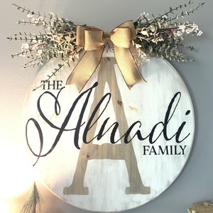 May include: A circular wooden family sign, painted white, featuring the name "Alnadi" in a stylish font. A large letter "A" is central to the design. The sign is decorated with a burlap bow and greenery, offering a rustic aesthetic.