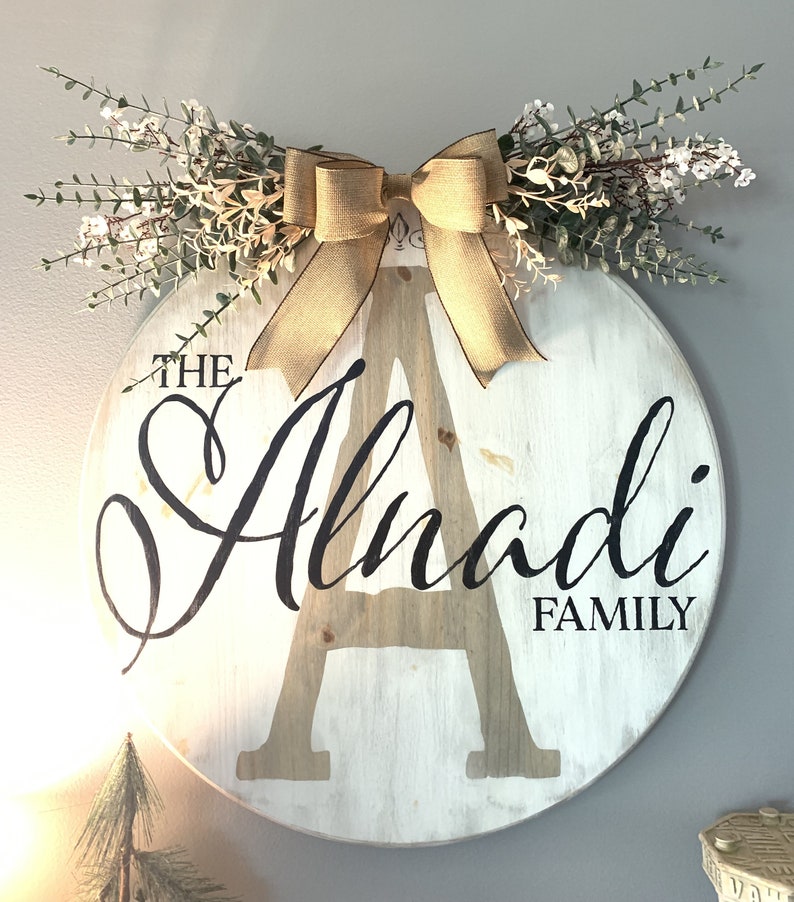 May include: A circular wooden family sign, painted white, featuring the name "Alnadi" in a stylish font. A large letter "A" is central to the design. The sign is decorated with a burlap bow and greenery, offering a rustic aesthetic.