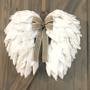 Pu&ograve; includere: Ali d'angelo in tessuto bianco con un fiocco in juta marrone, appese a una porta di legno.