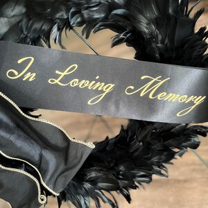 以下が含まれることがあります： 黒い羽根のリースで、金色の文字で「In Loving Memory」と書かれた黒いリボンが付いています。リースには金の花のアクセントと、金のトリムが付いた黒いサテン生地が付いています。