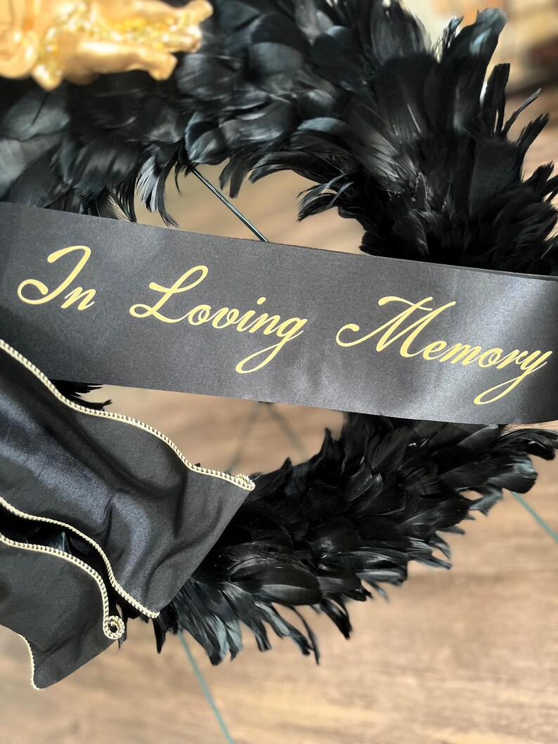 以下が含まれることがあります： 黒い羽根のリースで、金色の文字で「In Loving Memory」と書かれた黒いリボンが付いています。リースには金の花のアクセントと、金のトリムが付いた黒いサテン生地が付いています。