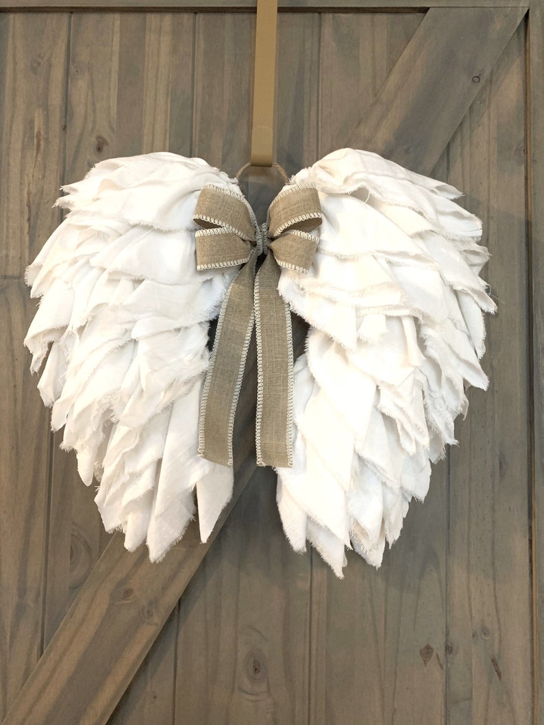 Pu&ograve; includere: Ali d'angelo in tessuto bianco con un fiocco in juta marrone, appese a una porta di legno.