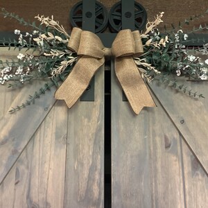 May include: A rustic barn door with a decorative burlap bow and floral arrangement. The door is grey with black hardware, and the bow is made of natural-toned burlap. The floral arrangement includes green and white elements.