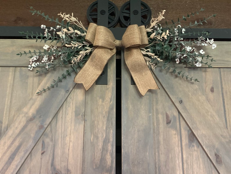 May include: A rustic barn door with a decorative burlap bow and floral arrangement. The door is grey with black hardware, and the bow is made of natural-toned burlap. The floral arrangement includes green and white elements.