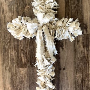 May include: A large, white, fabric cross with a bow in the center. The cross is made of strips of fabric that have been frayed and tied together. The cross is hanging on a wood wall.