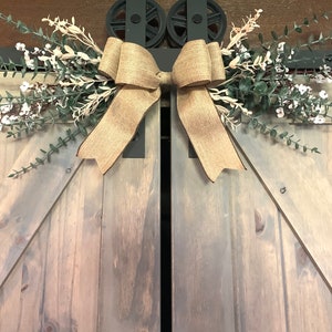 May include: A decorative arrangement for barn doors, featuring a large burlap bow and artificial greenery with white flowers. The piece is mounted above a set of grey wooden doors, creating a rustic, country-style aesthetic.