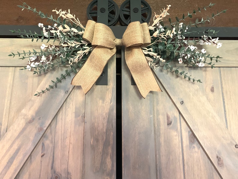 May include: A decorative arrangement for barn doors, featuring a large burlap bow and artificial greenery with white flowers. The piece is mounted above a set of grey wooden doors, creating a rustic, country-style aesthetic.