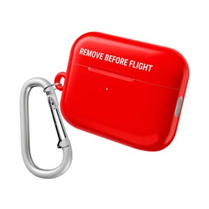 以下が含まれることがあります： 鮮やかな赤色のAirPodsケース。白い文字で「REMOVE BEFORE FLIGHT」と書かれています。シルバーのカラビナクリップが付いています。ワイヤレスイヤホンを保護し、持ち運ぶためのケースです。
