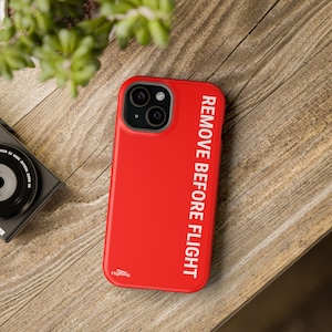 以下が含まれることがあります： 白い「REMOVE BEFORE FLIGHT」の文字が入った赤いiPhoneケース。ケースは光沢仕上げで、黒いカメラモジュールが付いています。木製の表面に置かれた携帯電話ケースです。