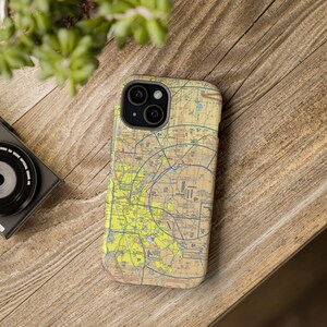 May include: A phone case featuring a detailed aviation map design in yellow, blue, and tan. The case is on a wooden surface with a camera and plant in the background. The case is designed to fit a smartphone.