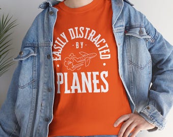 May include: Orange t-shirt with the text "EASILY DISTRACTED BY PLANES" in white, featuring an airplane graphic. The shirt is worn with a light blue denim jacket and blue jeans.