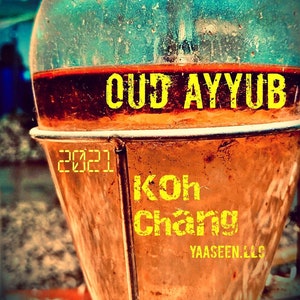 May include: A close-up of a rusty metal container with the words "OUD AYYUB KOH CHANG YAASEEN. LLC" printed on it. The container is filled with a dark liquid and has a clear glass lid with a small Arabic word written on it.
