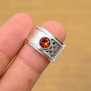 May include: A silver ring with a round, orange gemstone set in the center. The ring band has a textured pattern of small, repeating shapes. The ring is held by a finger.