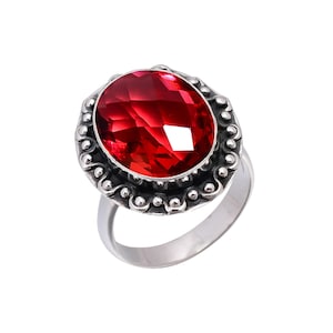 May include: A silver ring featuring a large, oval-shaped, faceted red gemstone set in a decorative bezel. The metal surrounding the stone has a dark, oxidized finish with a beaded edge detail, while the band itself is a smooth, polished silver.