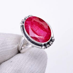May include: A silver ring featuring a large, oval-shaped, faceted deep pink gemstone set in a polished metal bezel. The ring band is split and decorated with small silver spheres on both sides of the central stone setting.