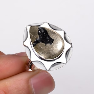 Puede incluir: Anillo de plata con una piedra de pirita en forma de pera, que muestra un patrón oscuro e irregular. La piedra está engastada en un bisel decorativo con un borde festoneado. La banda del anillo tiene un diseño retorcido.