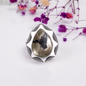 May include: A teardrop-shaped ring featuring a central metallic pyrite stone with a dark, irregular inclusion. The stone is encased in a polished silver-toned metal setting with a scalloped, petal-like border and smooth, dark gray inlay accents.