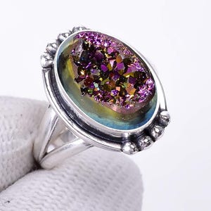 Könnte beinhalten: Silberring mit einem ovalen, irisierenden Druzy-Stein in Lila, Gold und Grün. Der Ring hat eine detaillierte Fassung mit kleinen, dekorativen Akzenten.