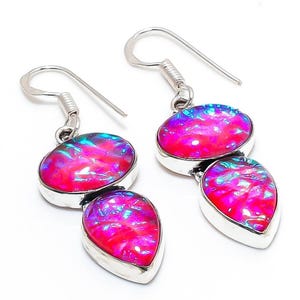 May include: A pair of drop earrings featuring two vibrant pink and iridescent blue opal-like stones set in polished silver. Each earring consists of an oval-shaped stone positioned above a teardrop-shaped stone, suspended from a silver hook wire.