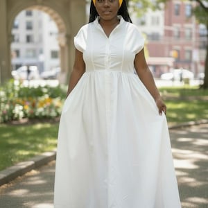 May include: A floor-length white dress with short sleeves, a button-down front, and a collar. The hem features a dark blue horizontal stripe and a yellow fringe trim. A bright yellow head wrap is worn on the head, matching the fringe detail.