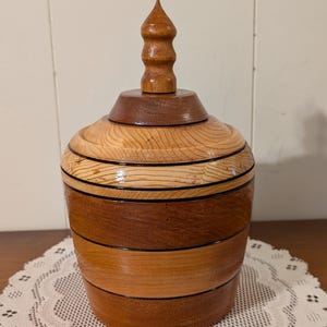 May include: A cylindrical wooden container with a removable lid featuring a pointed finial. The body is constructed from alternating horizontal bands of light and dark wood, separated by thin black rings, with a polished, smooth finish throughout.
