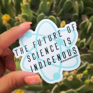 May include: A white sticker with a light blue atom design and the text "THE FUTURE OF SCIENCE IS INDIGENOUS." The sticker has a light blue border and is held in front of a blurred green background.