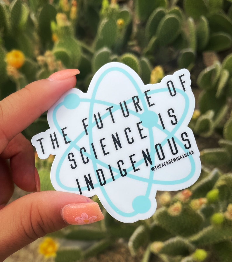 May include: A white sticker with a light blue atom design and the text "THE FUTURE OF SCIENCE IS INDIGENOUS." The sticker has a light blue border and is held in front of a blurred green background.