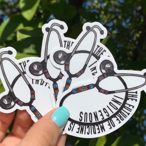 May include: Four white stickers with a black stethoscope design and the text "The future of medicine is indigenous" in a circle around the stethoscope. The stickers are stacked on top of each other.