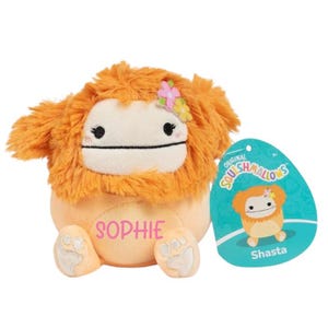 Peut inclure: Peluche orange et blanche avec une crini&egrave;re orange moelleuse et une fleur rose sur la t&ecirc;te. Le jouet a un visage blanc avec des yeux noirs et une bouche noire. Le jouet est assis sur une surface blanche et porte le nom "Sophie" brod&eacute; sur sa poitrine. Le jouet est un Squishmallow.