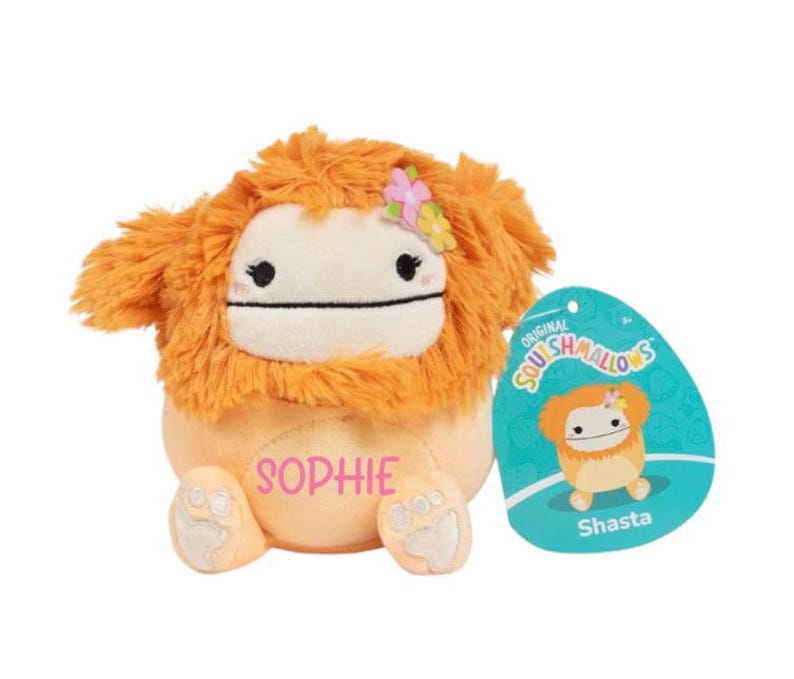 Peut inclure: Peluche orange et blanche avec une crini&egrave;re orange moelleuse et une fleur rose sur la t&ecirc;te. Le jouet a un visage blanc avec des yeux noirs et une bouche noire. Le jouet est assis sur une surface blanche et porte le nom "Sophie" brod&eacute; sur sa poitrine. Le jouet est un Squishmallow.