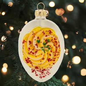 May include: A white glass Christmas ornament shaped like a plate with a swirl of yellow and pink frosting topped with red and green sprinkles.