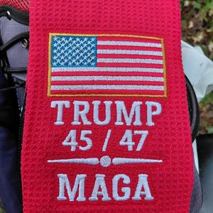 May include: A red golf towel with an embroidered American flag and the words "TRUMP 45 / 47 MAGA" in white. The towel is made of a textured material and is likely used for wiping golf clubs or hands.