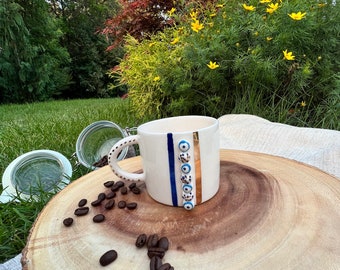 May include: White ceramic coffee mug with a blue and gold stripe design and blue evil eye charms. The mug is sitting on a wooden surface with coffee beans scattered around it.