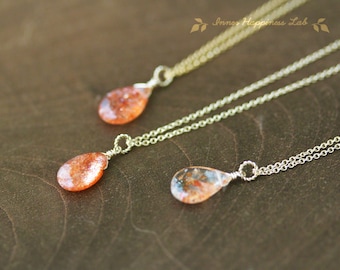 May include: Three gold necklaces with sunstone pendants. Each pendant is a teardrop shape with a different pattern of inclusions. The necklaces are layered on a brown wooden surface.