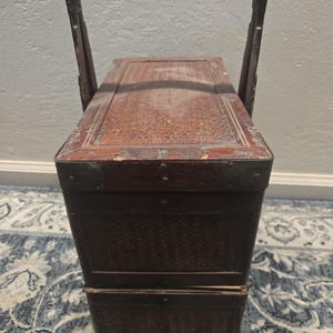 Könnte beinhalten: Ein rechteckiger, dunkelbrauner Vintage-Korb mit gewebter Oberfläche, einem Klappdeckel und einem gebogenen Griff. Der Korb hat Metallecken und ein gewebtes Muster an den Seiten und oben. Der Korb steht auf einem blau-weiß gemusterten Teppich.