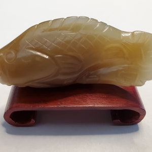 May include: A carved jade fish figurine in a light brown color, resting on a small, dark red wooden stand.