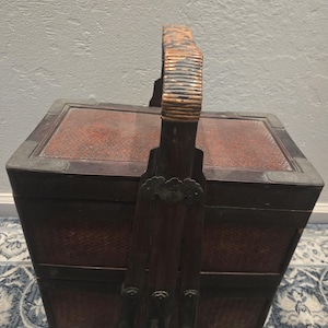 May include: A vintage, two-tiered, dark brown woven basket with a hinged lid and a sturdy handle. The basket features a rectangular shape with decorative metal accents and a reddish-brown interior. The handle is wrapped with a lighter-colored material.