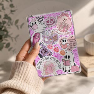 Może przedstawiać: Przezroczyste etui na tablet z różowym tłem w pajęczynę i różnymi naklejkami, w tym czytającymi duchami, szkieletem, dyniami, trumną i księgą zaklęć. Tekst zawiera napisy "hot ghouls read" oraz "one more chapter".