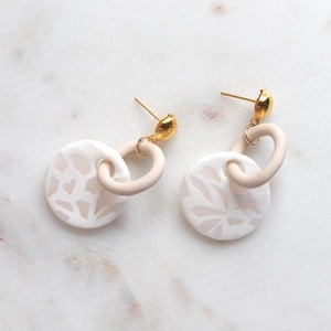 May include: A pair of white ceramic earrings with a gold-toned metal loop and a white floral pattern. The earrings are on a white marble surface.