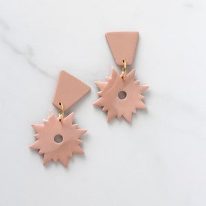 May include: A pair of pink clay earrings with a starburst shape and a triangle shape. The earrings are connected by a gold ring.