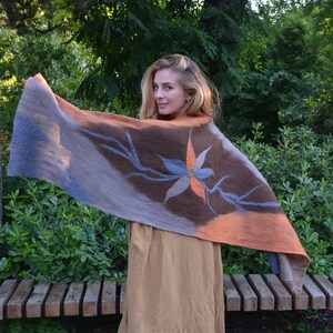 Könnte beinhalten: Eine Frau hält einen Filzschal mit floralem Design hoch. Der Schal zeigt Brauntöne, Blau und Orange. Die Frau trägt ein beiges Kleid und braune Schuhe, vor einer Parkbank und Grün.