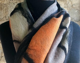 May include: A felted wool scarf in shades of gray, orange, and black, draped on a black mannequin. The scarf features a geometric design with bold color blocks and a soft, textured appearance. The scarf is a stylish accessory.