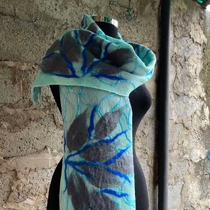 Könnte beinhalten: Hellblauer Filzschal mit einem Blattmuster in Grau und Blau. Der Schal ist über einer schwarzen Schaufensterpuppe drapiert und zeigt die komplizierten Details der Filzwolle. Das Blattmuster wird mit blauen Nähten erzeugt.