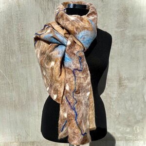 Könnte beinhalten: Ein Filzschal aus Wolle in Braun-, Blau- und Weißtönen, drapiert auf einer schwarzen Schaufensterpuppe. Der Schal hat ein strukturiertes Design mit blauen Nähten und weißen Tupfen. Ein Winteraccessoire.