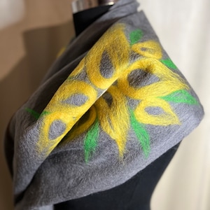 Könnte beinhalten: Grauer Schal mit gelbem Blumenmuster und grünen Akzenten. Der Schal ist über einer Schaufensterpuppe drapiert, was die weiche Textur und die leuchtenden Farben des handgefertigten Accessoires hervorhebt. Ein stilvolles Accessoire.