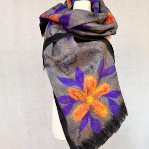 Könnte beinhalten: Grauer Filzschal mit großem orange-violettem Blumenmuster. Der Schal hat einen schwarzen Rand und ist über einer weißen Schaufensterpuppe drapiert. Der Schal hat auch orangefarbene und violette geometrische Muster.