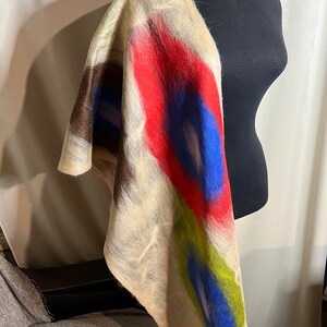 Könnte beinhalten: Ein beigefarbener Filzschal aus Wolle mit leuchtenden Farbakzenten. Das Design zeigt kräftige Striche in Rot, Blau und Grün, die einen auffälligen visuellen Kontrast erzeugen. Der Schal ist über einer schwarzen Schaufensterpuppe drapiert, um seine Form und Textur zu präsentieren.